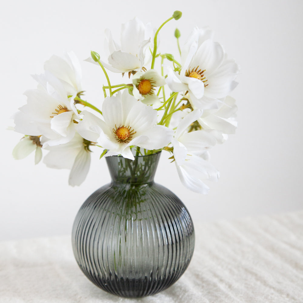 Artificial white cosmos flowers in a small dark glass vase.
