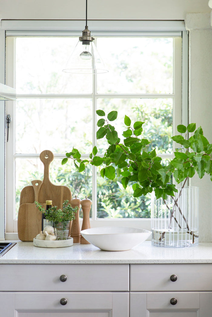 Styled kitchen benchtop with a vase with green leaves, wooden chopping boards and a fruit bowl
