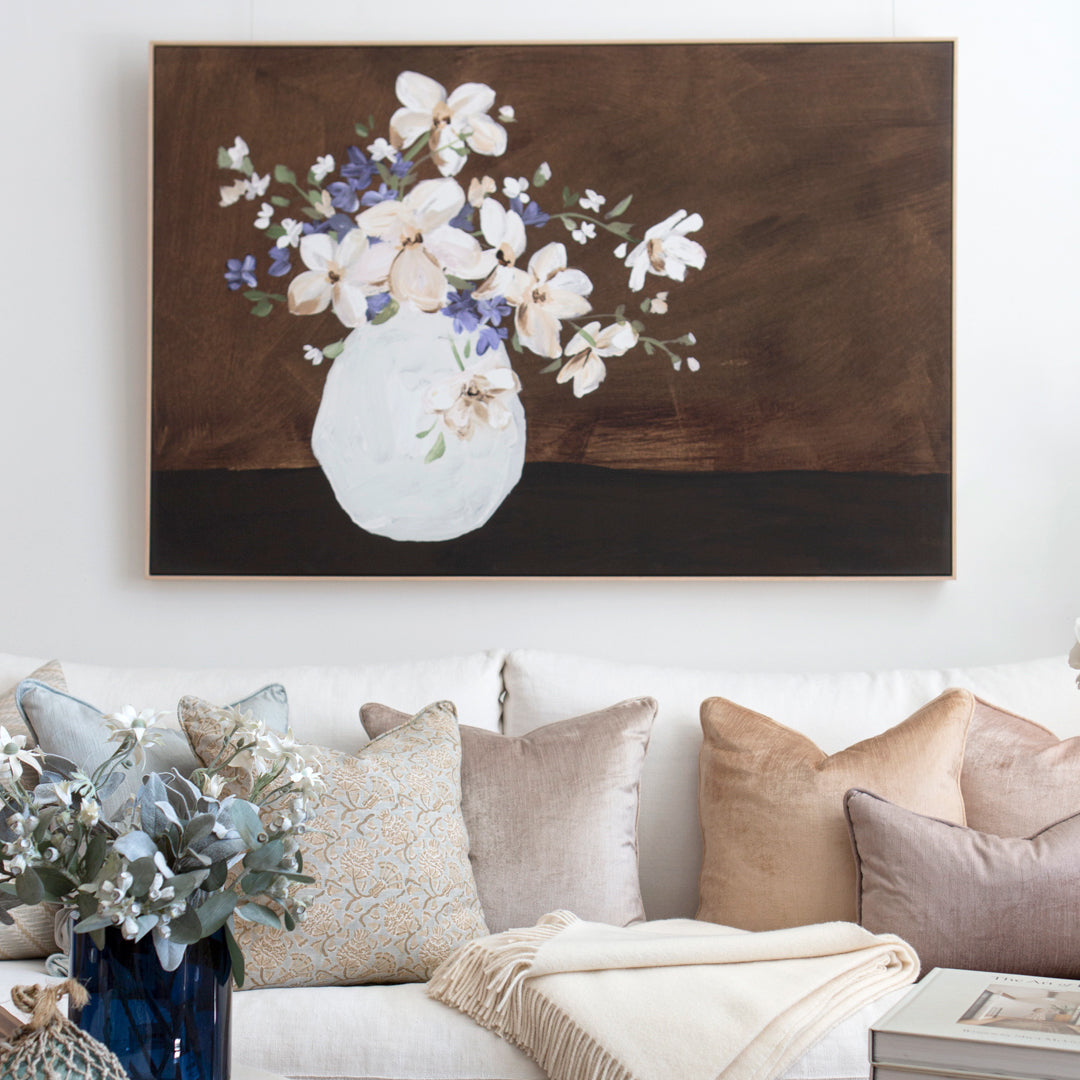 Rectangular canvas wall art print of a painting. Artwork features a vase of soft blush and blue flowers off to the left side with a brown painterly background. Artwork featured above white sofa with cushions.