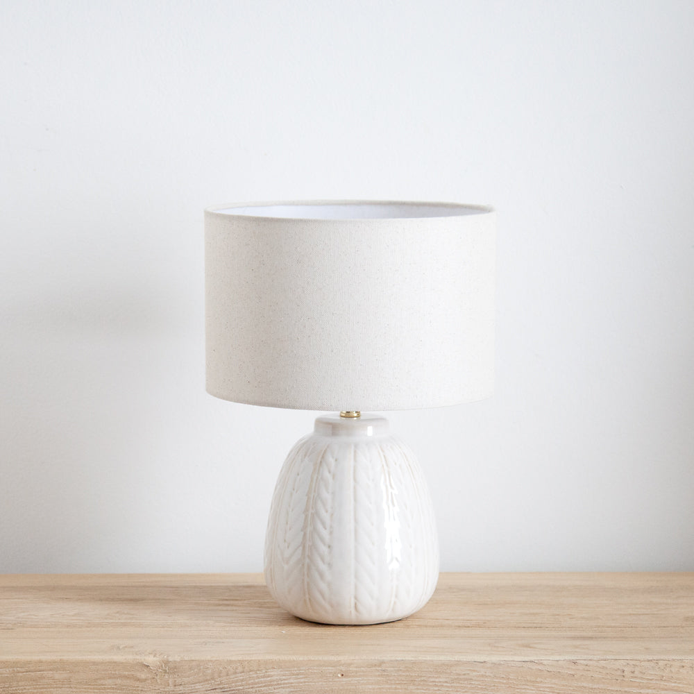 Small white ceramic lamp with linen shade. Displayed on wooden table.