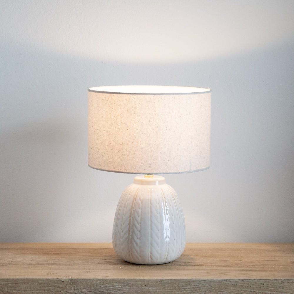 Small white ceramic lamp with linen shade. Shown on a wooden table and turned on. 