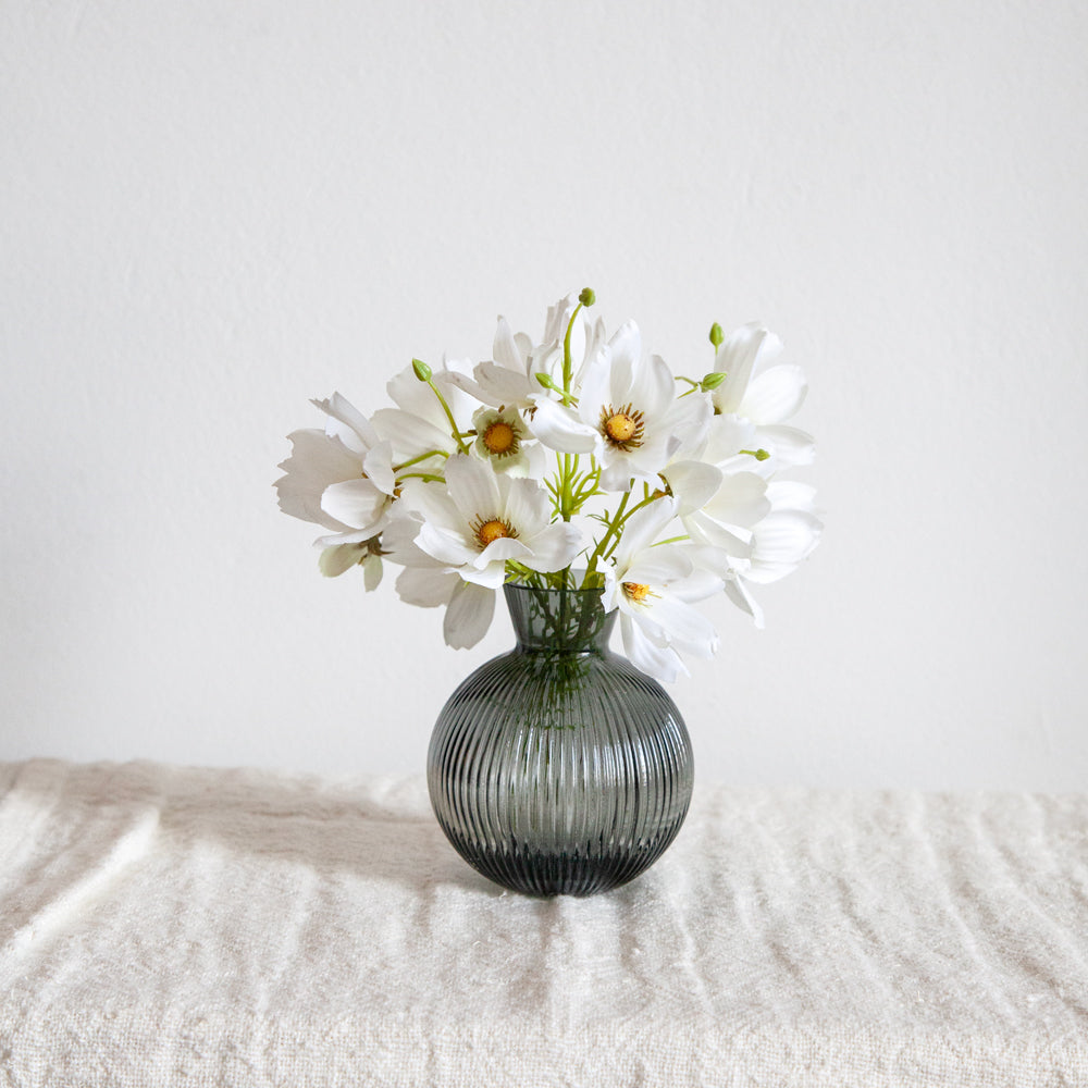 Artificial white cosmos flowers in a small dark glass vase.