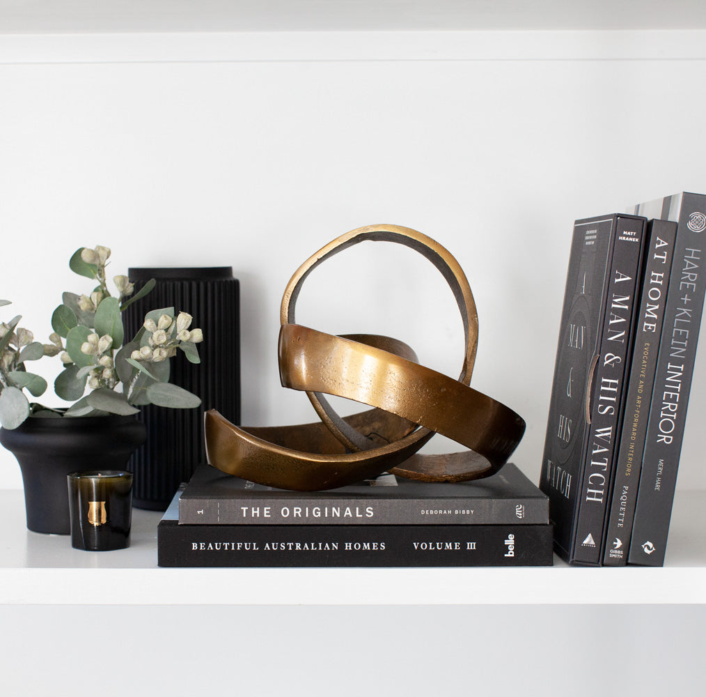 Freeform brass sculpture styled on shelf with black books and vases.