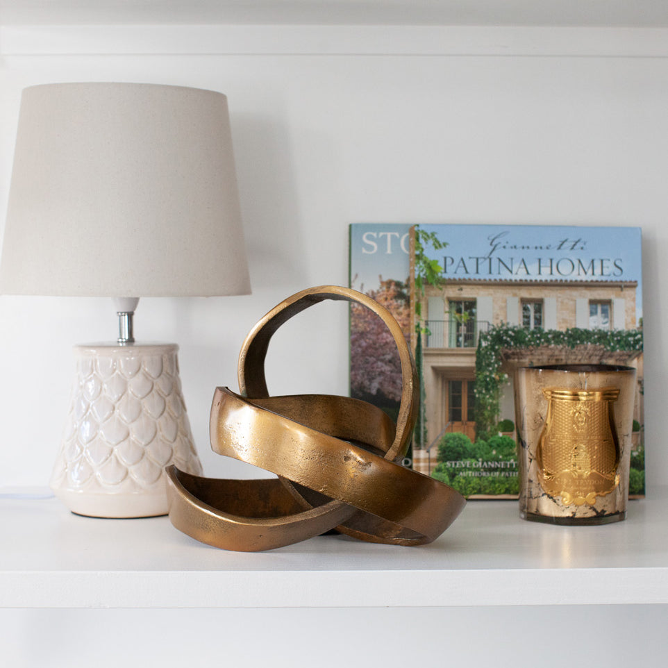 Freeform brass sculpture styled on shelf with a lamp, books and a candle..